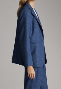 Femme portant un blazer bleu cintré et un pantalon assorti, debout devant un fond gris uni, vue de côté.