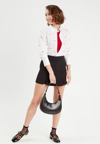 Chemise blanche à manches longues avec des appliqués en forme de cœur rouge, haut rouge, jupe noire, chaussettes noires à motifs et sac à main en cuir noir à effet crocodile.
