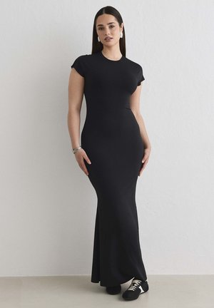 Kobieta o długich ciemnych włosach ma na sobie dopasowaną czarną suknię maxi z krótkimi rękawami typu casquette oraz czarne trampki z białymi sznurowadłami, stoi oparta o białą ścianę.