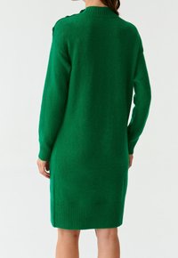Robe en tricot verte avec une coupe décontractée, des poignets et un ourlet côtelés, et un détail de boutons aux épaules. Texture douce, longueur sous le genou.