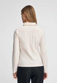 Beżowa koszulka polo z długim rękawem, kołnierzykiem, miękkim materiałem, bocznymi rozcięciami i luźnym krojem, noszona z czarnymi spodniami. Widok od tyłu.