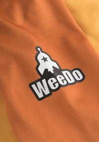 Oranssi kangas esittää "WeeDo"-logoa mustavalkoisena, jossa on kellomuoto tähti, teksturoidulla taustalla.