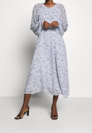 Femme portant une robe midi lavande claire avec un imprimé noir éclaboussé et des manches longues bouffantes, associée à des talons ouverts noirs.