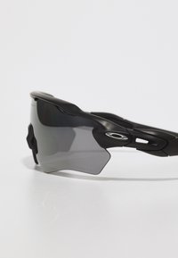 Czarne sportowe okulary przeciwsłoneczne z szarymi przyciemnionymi soczewkami, aerodynamiczny kształt oraz gumowe uchwyty dla komfortu, z emblematem po boku.