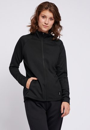 Jonge vrouw met schouderlang bruin haar, draagt een zwart jack met rits en bijpassende zwarte broek, staand met één hand in de zak.