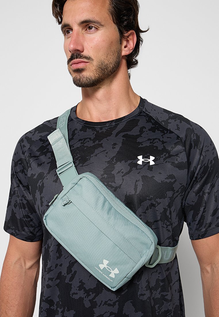 Hombre con camiseta Under Armour de camuflaje negro y bolso cruzado de Under Armour de color verde claro sobre el pecho.