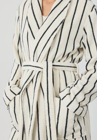 Krém színű, fekete függőleges csíkos fürdőköpeny, puha törölközőanyagból készült. Kialakítása sálgalléros, övvel és oldalzsebbel rendelkezik.