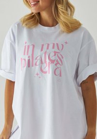 Biała, oversize'owa koszulka z krótkimi, podwiniętymi rękawami, z różowym napisem "w mojej erze pilatesu", ozdobiona małymi różowymi gwiazdkami.