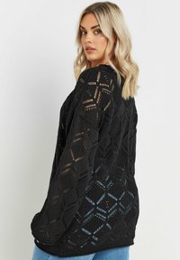 Svart, öppet stickad tröja med ett geometriskt diamantmönster, långa ärmar och en avslappnad passform. Designen visar upp intrikat vävda detaljer.