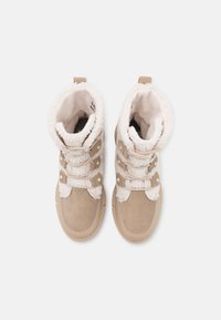 Suede beige støvler med hvid pelsforing, tre dekorative remme, rund tå og gummisåler. Accenter inkluderer metalknapper og sydetaljer.