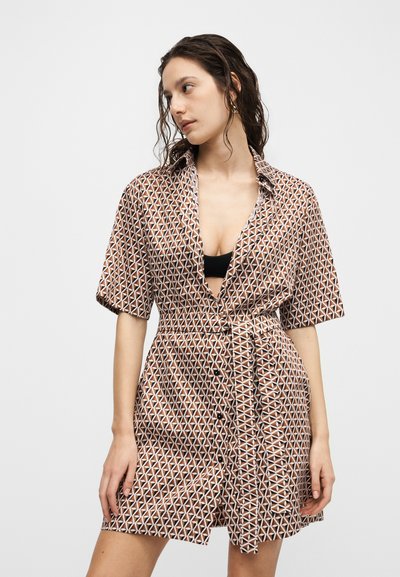 Γυναίκα με καφέ φόρεμα με γεωμετρικό σχέδιο και ζώνη, με κοντά μανίκια και μαύρο τοπ από κάτω, κοιτάζει προς τα πλάγια.