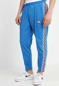 Muž v modrých teplácích značky Nike s bílými, červenými a modrými pruhy na boku a bílých teniskách Nike s černými logy swoosh.