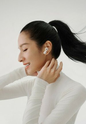 Wireless earbuds in a white color worn by a person with long dark hair, who is dressed in a fitted white long-sleeve top, smiling.