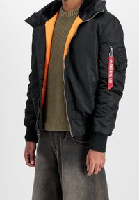 Blouson aviateur noir avec doublure orange, fermeture éclair, poignets en tricot et poche à patch sur la manche gauche avec une sangle rouge.