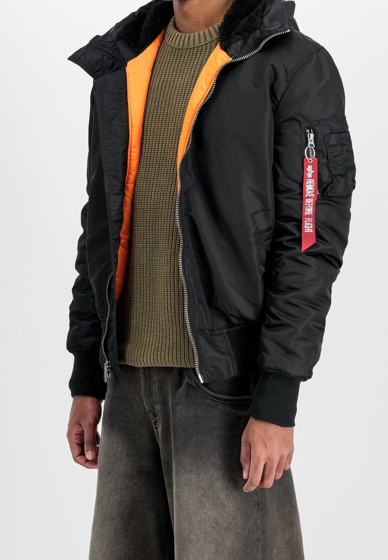 Blouson aviateur noir avec doublure orange, fermeture éclair, poignets en tricot et poche à patch sur la manche gauche avec une sangle rouge.