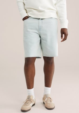 Osoba stojaca s jednou rukou v vrecku, oblečená v svetlomodrých riflových šortkách, bielej mikine, bielych ponožkách a béžových teniskách.