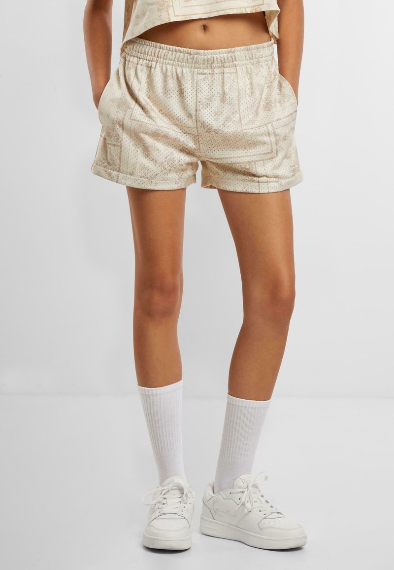 Beige gemusterte Shorts mit elastischem Taillenbund, seitlichen Taschen und lockerer Passform, kombiniert mit weißen knielangen Socken und Sneakers.