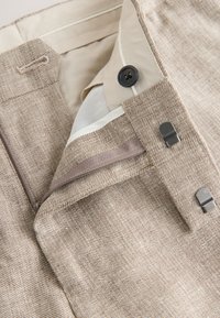 Close-up of beige textured pants waistband showing button, hook-and-bar fasteners, and inner lining details.