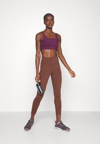 Violeta sporta krūšturis un brūnas augstā jostasvieta legingi, izgatavoti no elastīga auduma. Modelis tur melnu ūdens pudeli un nēsā pelēkus sporta apavus.