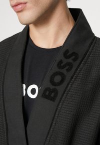 Črna teksturirana halja z širokim ovratnikom, ki vključuje puhasto izbodeno oznako "BOSS" na levi strani, oblečena čez črno majico.