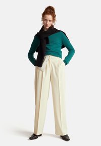 Zelenomodrý pletený svetr, černý šátek přes ramena, krémové skládací kalhoty a černé boty. Modelka stojí s rukama v kapsách.