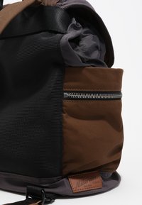Rjava in črna tkanina nahrbtnik ima mrežno stransko ploščo, žep na zadrgo in teksturirano usnjeno oznako blagovne znamke.