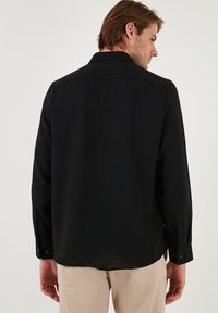 Camisa negra de manga larga, fabricada en tejido ligero, con puños abotonados y cuello clásico. Textura suave, corte entallado y diseño limpio en la parte de atrás.