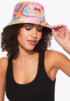 Vrouw met krullend haar die een kleurrijke vissershoed en een zwart mouwloos bovenstuk draagt, terwijl ze de rand van de hoed zachtjes vasthoudt met haar rechterhand.