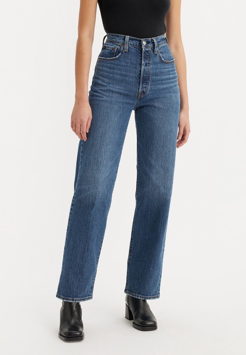 Femme portant un jean bleu taille haute, coupe droite, un top noir sans manches et des bottines noires à talons sur fond blanc.