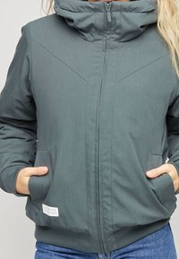 Graue Kapuzenjacke mit Reißverschluss vorne, schrägen Nähten und Seitentaschen. Hergestellt aus glattem, strukturiertem Stoff, taillierte Passform.