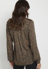 Blouse florale verte avec manches longues, détail plissé au dos et tissu texturé orné de motifs floraux orange et bleus.