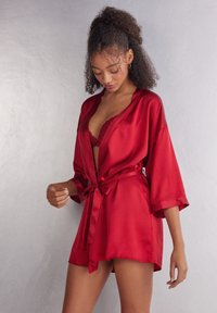 Röd satinrosett med knytmidja, 3/4-långa ärmar och en mjuk textur, lager på lager över en spets-bustier. Slät, glansig yta framhäver designen.