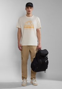 Beige grafisk t-shirt med orange bjergdesign, khaki cargo bukser og sort rygsæk. Hvide sneakers fuldender outfittet.