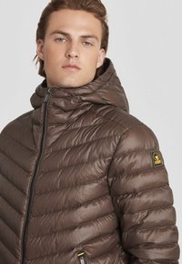 Giacca imbottita marrone con colletto alto, chiusura con zip e cuciture orizzontali. Include una patch con il logo sulla manica sinistra.