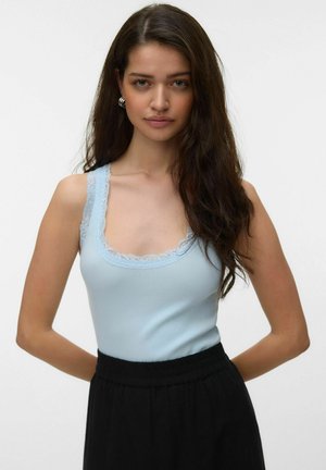 Joven mujer con cabello largo y oscuro, vestida con una camiseta sin mangas azul claro con encaje y pantalones negros de cintura alta, de pie con las manos detrás de la espalda.