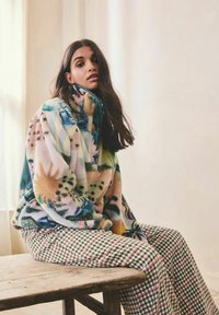 Tânără cu păr lung și închis la culoare, stând pe o bancă de lemn, purtând o jachetă fleece colorată cu model și pantaloni verzi în carouri, pe fundalul unui perete simplu.