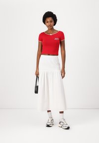 Camiseta roja corta con mangas cortas y borde blanco, combinada con una falda blanca de largo hasta los tobillos y con fruncido. Bolso negro y zapatillas.