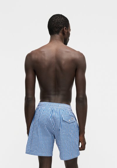 Άνδρας που φαίνεται από πίσω, φοράει μπλε και λευκά καρό σορτς, στέκεται μπροστά σε ένα απλό ανοιχτό γκρι φόντο.