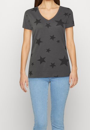 Kobieta ubrana w antracytowy T-shirt z dekoltem w serek z rozsianym czarnym wzorem gwiazdek i jasnoniebieskie dżinsy, stoi na tle jednolitego tła.