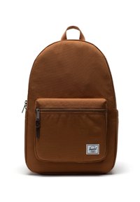 Brun tygryggsäck med rundad form, zip-magnetisk frontficka och en vit etikettaccent. Utmärks av ett handtag på toppen och justerbara axelremmar.