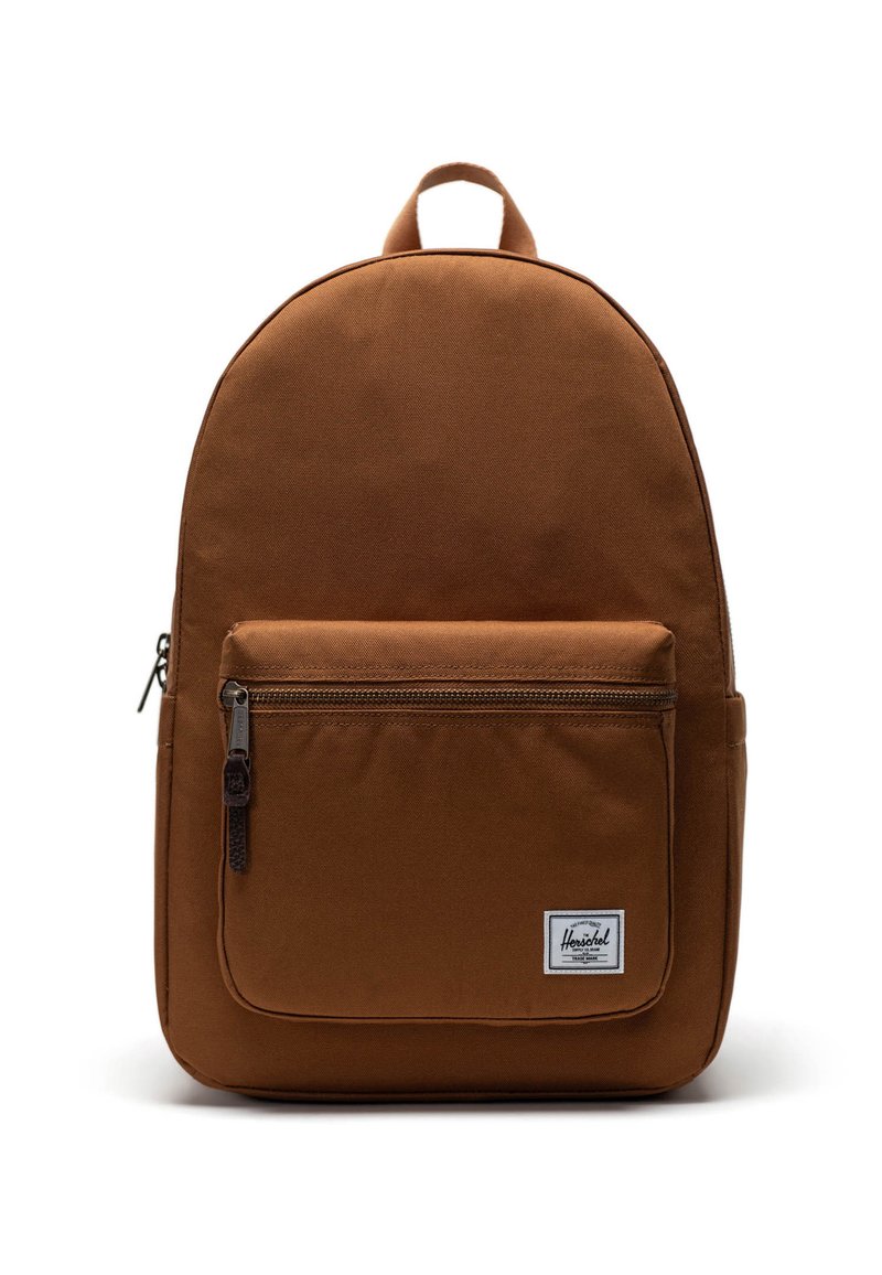 Brun tygryggsäck med rundad form, zip-magnetisk frontficka och en vit etikettaccent. Utmärks av ett handtag på toppen och justerbara axelremmar.