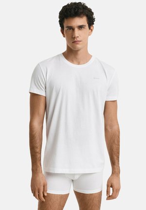 Homme portant un T-shirt blanc à manches courtes et un boxer blanc, se tenant devant un fond gris clair uni.