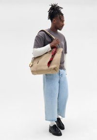 Bolso tote de lona beige con una bufanda roja de estampado atada al asa, que incluye un zipper y una correa para el hombro. El modelo lleva una camiseta gris y jeans claros.