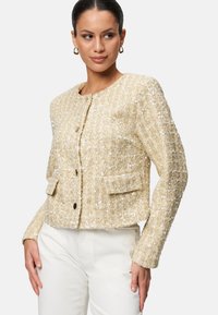 Giacca beige con paillettes, dalla finitura testurizzata, scollo rotondo, chiusura a bottoni e due tasche frontali, progettata con una silhouette accorciata.