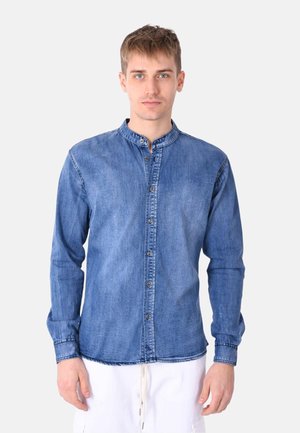 Camicia in denim di blu medio, con colletto alla coreana, chiusura con bottoni sul davanti e maniche lunghe con polsini. Texture di colore chiaro con cuciture visibili.