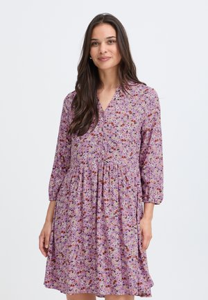 Kvinde med langt mørkt hår iført en lilla blomsterkjole til knæet med trekvartærmer og en rynket talje, stående mod en ensfarvet baggrund.