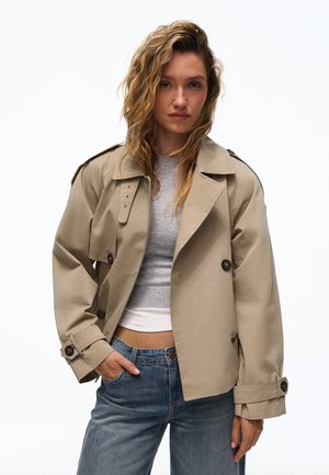Femme aux cheveux ondulés portant un trench beige par-dessus un top gris court et un jean bleu, debout avec une main dans la poche sur fond blanc.