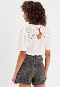Blouse blanche avec un dos en dentelle crochetée et des manches courtes bouffantes, associée à un short en denim gris foncé à taille haute, retroussé.