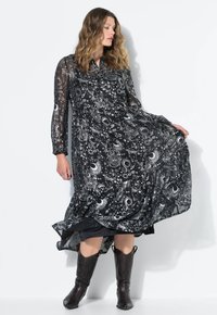 Vestito nero trasparente con intricati motivi bianchi di lune e disegni celestiali. Maniche lunghe, davanti con bottoni e orlo asimmetrico. Abbinato a stivali neri.