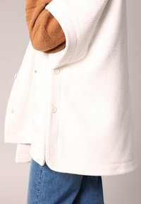 Weißer strukturierter Mantel mit kragen, ausgestattet mit einer Knopfleiste und seitlichen Taschen. Kombiniert mit blauen Jeans.